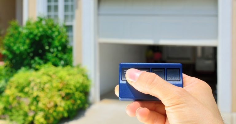 A hand is holding up a garage door opener remote to their garage door. The garage door in the background is about halfway open.