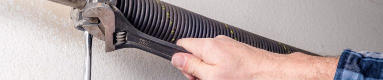 A pair of hands is using a wrench to perform some household maintenance on a large garage door spring.