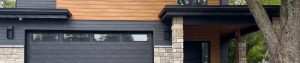 A view from the driveway of a modern home with a black garage door, a stone and wood facade, and large windows.